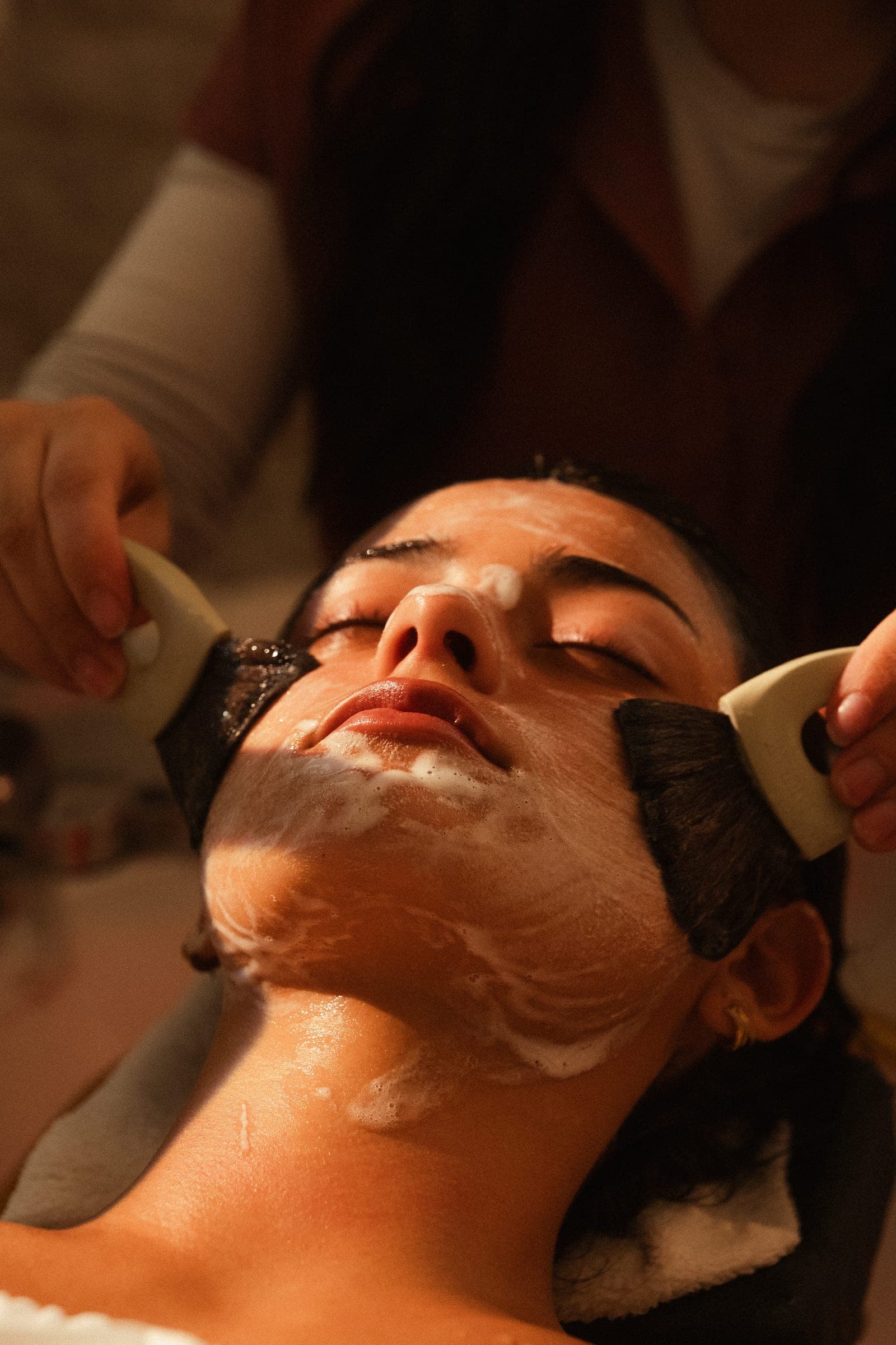 Two brushes working a cleansing foam across a guest's face during a facial ritual.